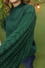 Load image into Gallery viewer, Model is wearing a green sweater in front of a festive background. 
