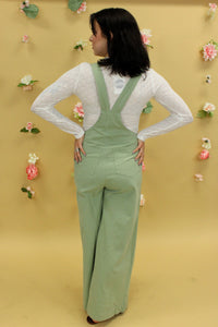 Model is wearing a light green overall with a white long sleeve top. 